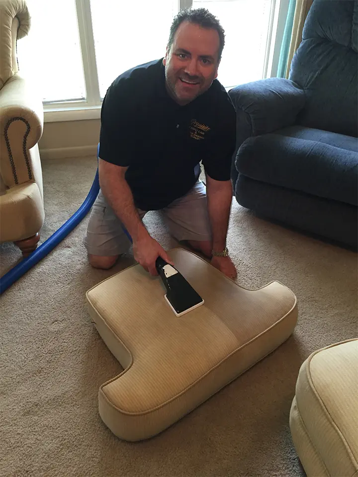 A man kneeling down on the floor cleaning a couch.