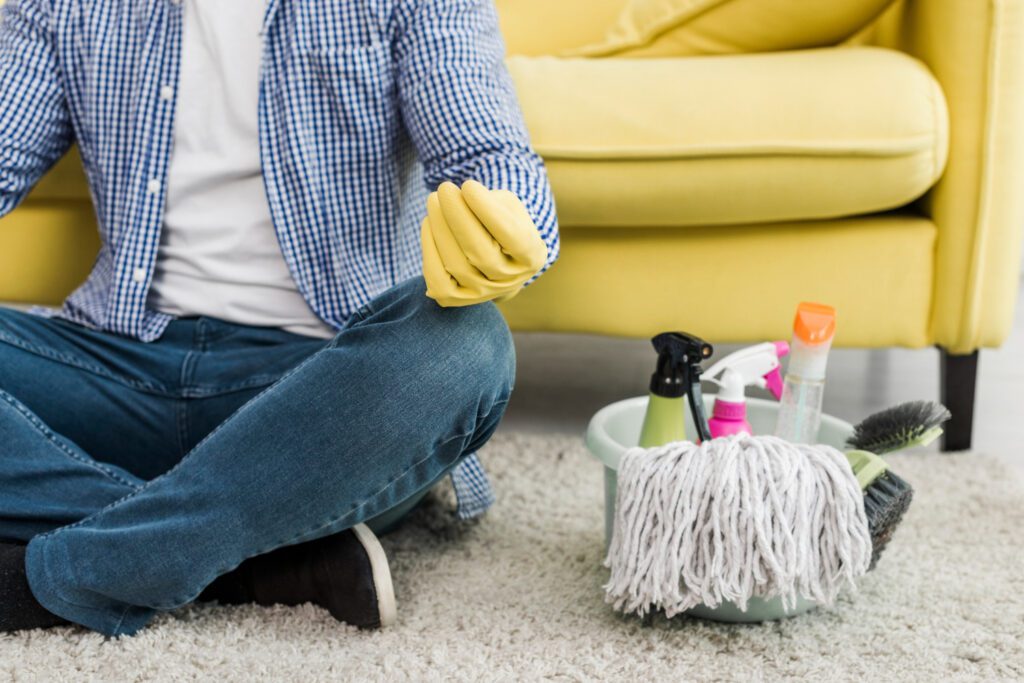 Person wearing gloves cleaning carpet with mop and cleaning supplies.